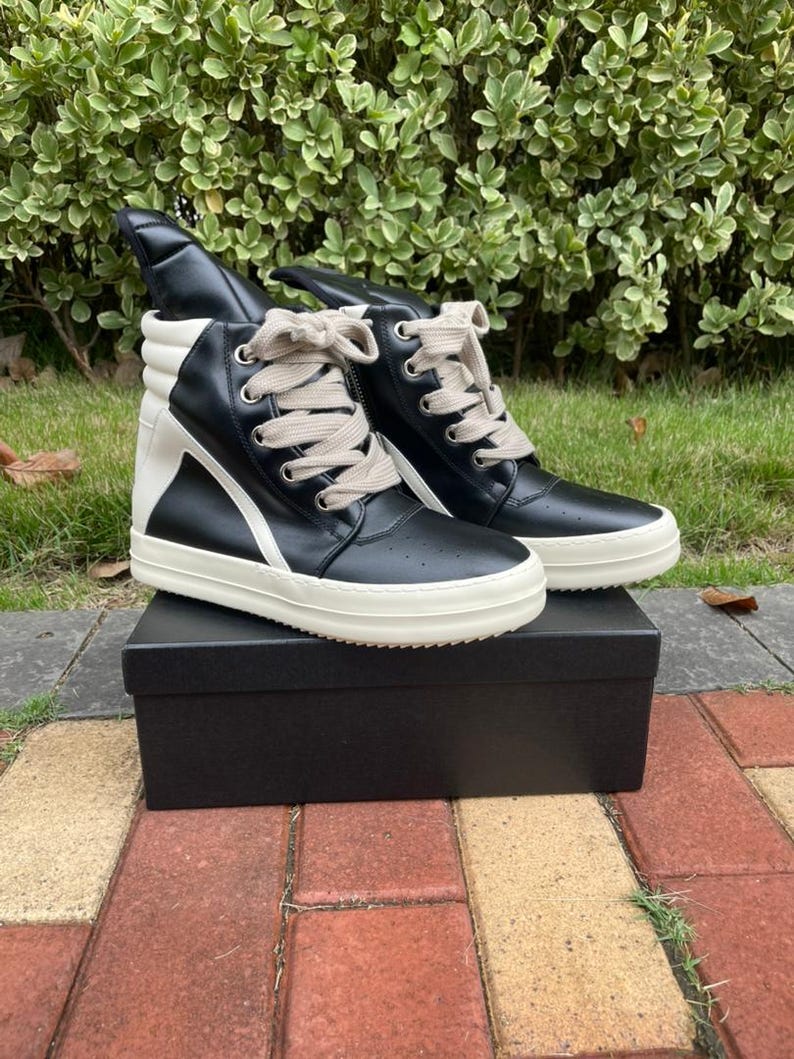 May include: A pair of high-top sneakers in black leather with white accents and a white sole. The sneakers have a unique design with a high tongue and are laced with beige laces. They are displayed on a black box.