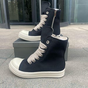May include: A pair of black high-top canvas sneakers with white soles and beige laces. The sneakers have a zipper on the side and are sitting on a gray box.