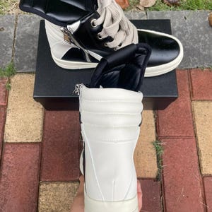 May include: High-top sneakers in black and white leather. The shoes feature a side zipper, padded tongue, and thick, off-white soles. The laces are a light beige color. The sneakers are on top of a black box.