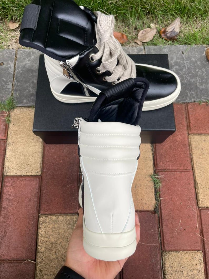 May include: High-top sneakers in black and white leather. The shoes feature a side zipper, padded tongue, and thick, off-white soles. The laces are a light beige color. The sneakers are on top of a black box.