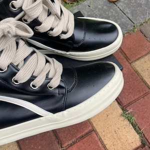 May include: Black high-top sneakers with cream-colored soles and laces. The sneakers have a perforated design on the upper and a white stripe detail. The laces are thick and the eyelets are silver.
