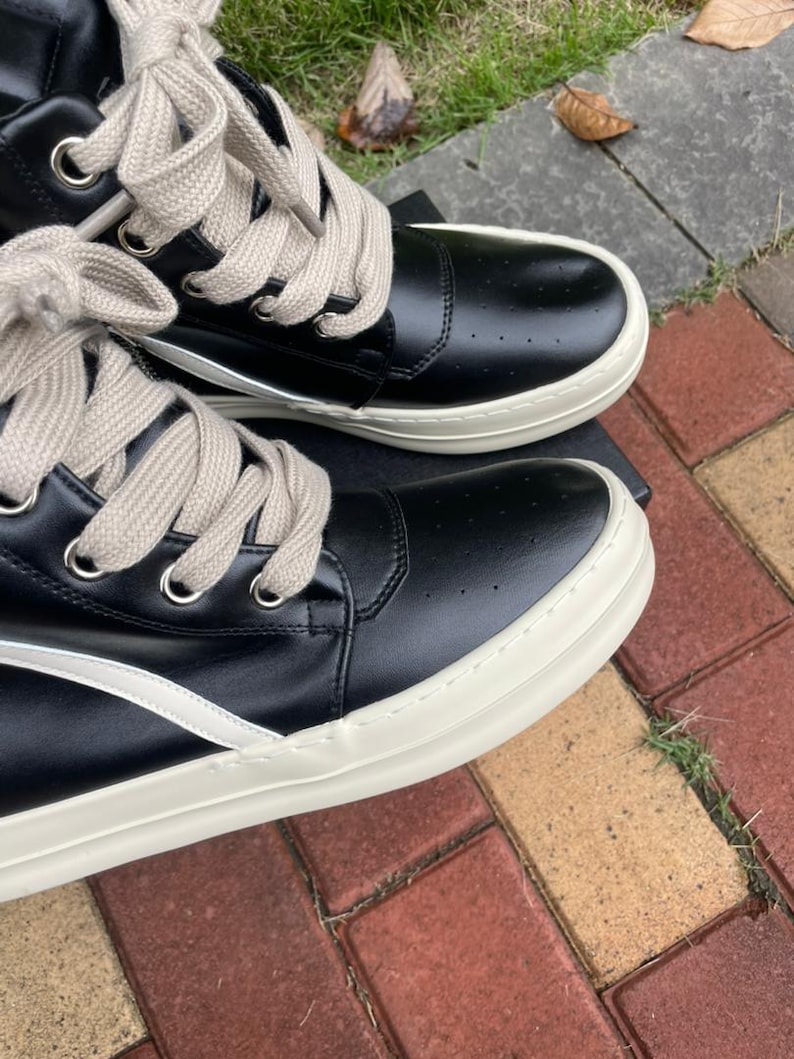 May include: Black high-top sneakers with cream-colored soles and laces. The sneakers have a perforated design on the upper and a white stripe detail. The laces are thick and the eyelets are silver.