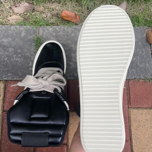 May include: Black high-top sneaker with a white sole and light-colored laces. The shoe features a padded ankle support and a textured sole. The sneaker is shown next to its sole, which is white with horizontal grooves.