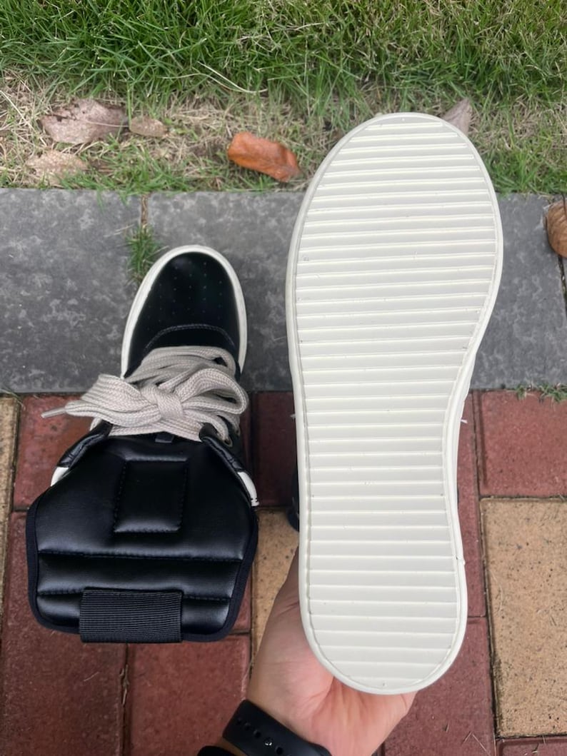 May include: Black high-top sneaker with a white sole and light-colored laces. The shoe features a padded ankle support and a textured sole. The sneaker is shown next to its sole, which is white with horizontal grooves.