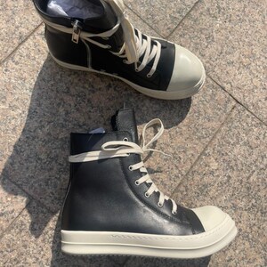 May include: A pair of high-top sneakers featuring black leather uppers, off-white rubber toe caps, and thick off-white rubber soles. The shoes include off-white laces, silver-toned metal eyelets, and a silver-toned side zipper on the inner ankle.