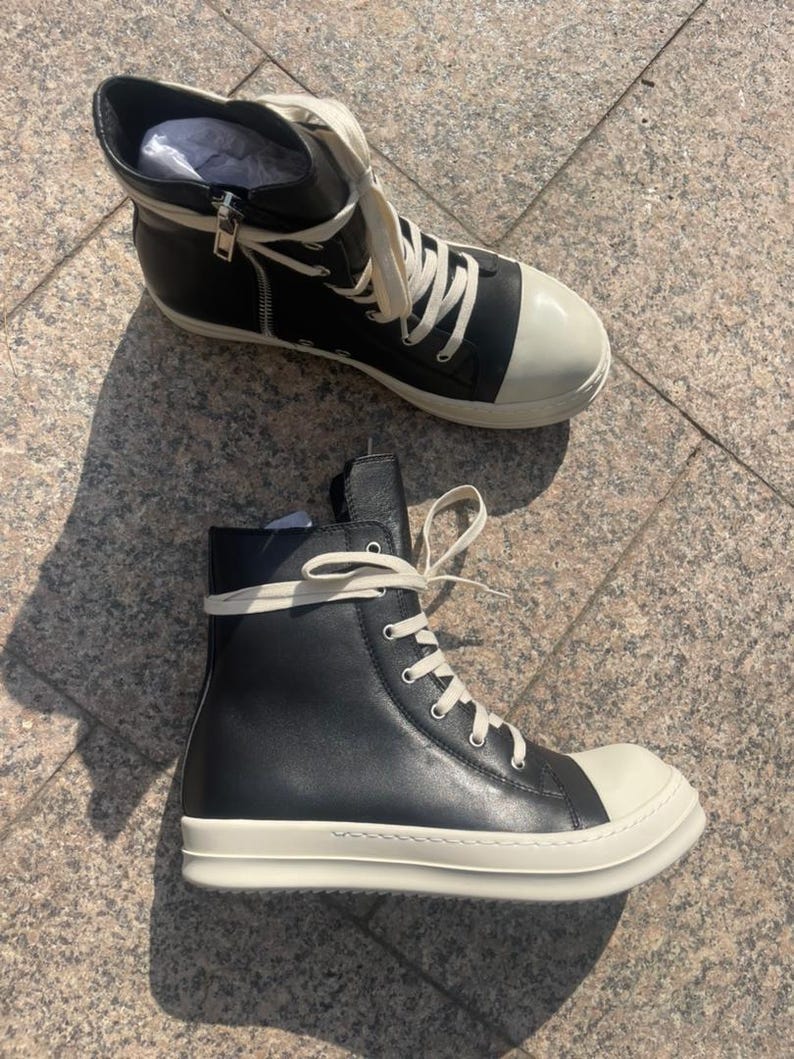 May include: A pair of high-top sneakers featuring black leather uppers, off-white rubber toe caps, and thick off-white rubber soles. The shoes include off-white laces, silver-toned metal eyelets, and a silver-toned side zipper on the inner ankle.