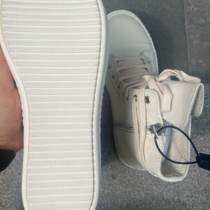 May include: Off-white high-top sneakers with a side zipper and lace-up closure. The sole is white with a ridged design. The shoe has a tag attached. The sneakers are made of a smooth material.