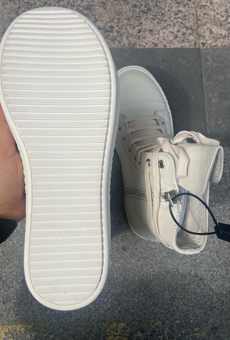 May include: Off-white high-top sneakers with a side zipper and lace-up closure. The sole is white with a ridged design. The shoe has a tag attached. The sneakers are made of a smooth material.