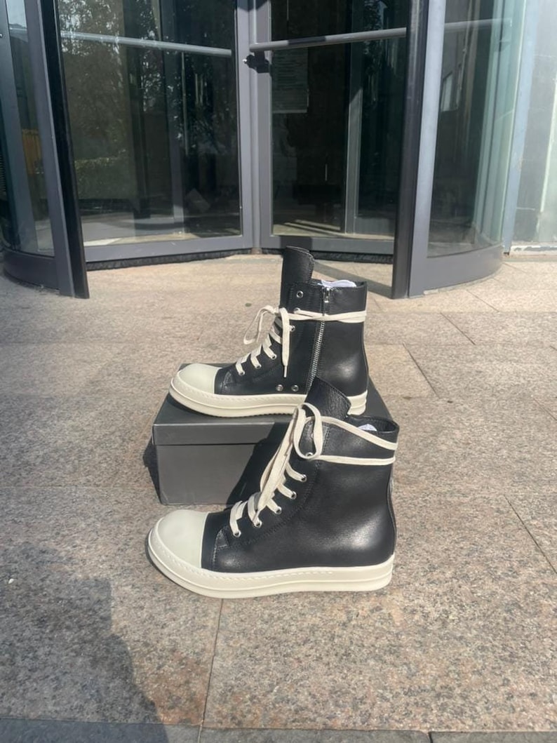 May include: A pair of black high-top sneakers featuring a smooth leather upper, a rounded off-white rubber toe cap, and a thick off-white rubber sole. The sneakers include long off-white laces and a silver-toned side zipper on the inner ankle.