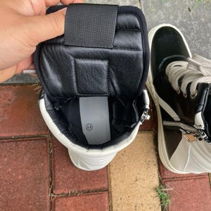 May include: High-top sneakers in black and white leather. The sneakers have a zipper closure and a white sole. The interior is black with a padded collar and a black strap. The size 44 is visible inside the shoe.