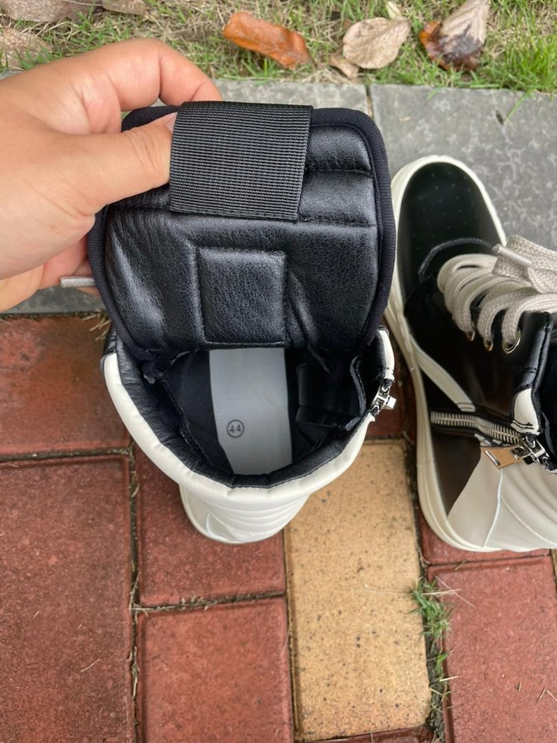 May include: High-top sneakers in black and white leather. The sneakers have a zipper closure and a white sole. The interior is black with a padded collar and a black strap. The size 44 is visible inside the shoe.