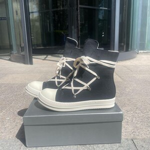 May include: A pair of black high-top canvas trainers with white laces and a white rubber sole. The shoes have a unique criss-cross lacing pattern and are sitting in a grey box.