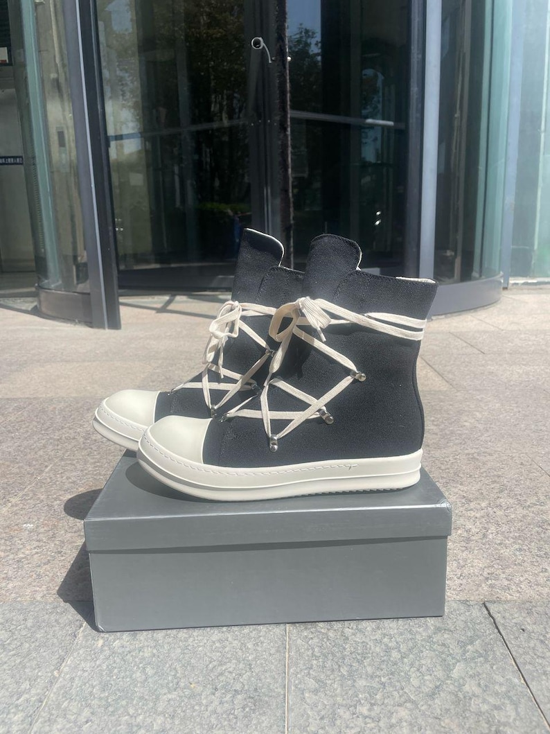 May include: A pair of black high-top canvas trainers with white laces and a white rubber sole. The shoes have a unique criss-cross lacing pattern and are sitting in a grey box.