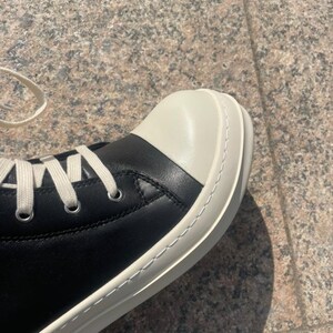 May include: A high-top sneaker featuring a smooth black leather upper, a prominent off-white rounded rubber toe cap, and a thick off-white rubber sole with visible stitching. The shoe includes silver-toned metal eyelets and flat off-white laces.