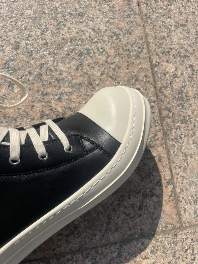 May include: A high-top sneaker featuring a smooth black leather upper, a prominent off-white rounded rubber toe cap, and a thick off-white rubber sole with visible stitching. The shoe includes silver-toned metal eyelets and flat off-white laces.