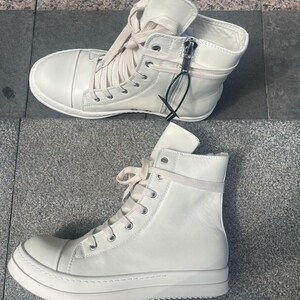 May include: Two pairs of off-white high-top sneakers. The sneakers have laces, zippers, and a thick sole. The shoes are made of a smooth material and are designed for casual wear.