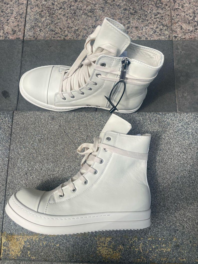 May include: Two pairs of off-white high-top sneakers. The sneakers have laces, zippers, and a thick sole. The shoes are made of a smooth material and are designed for casual wear.