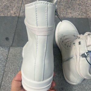 May include: White high-top sneakers with a side zipper and laces. The sneakers have a minimalist design with a smooth, clean surface and a thick sole. The shoes are made of a leather-like material.