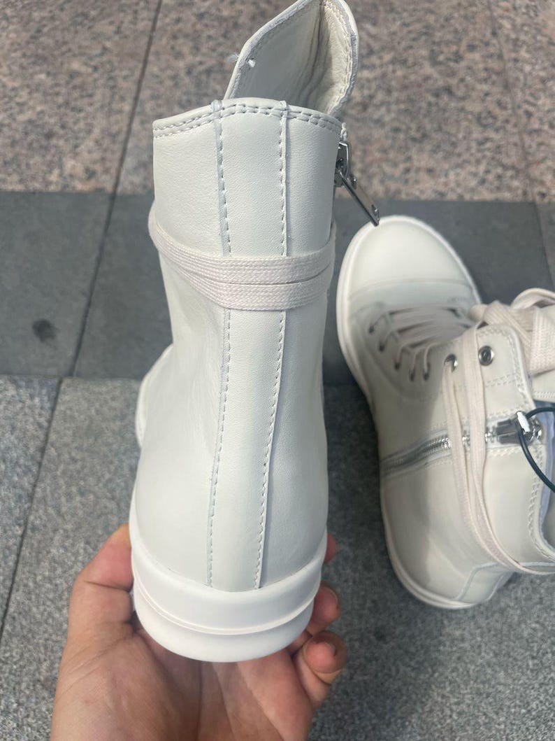 May include: White high-top sneakers with a side zipper and laces. The sneakers have a minimalist design with a smooth, clean surface and a thick sole. The shoes are made of a leather-like material.