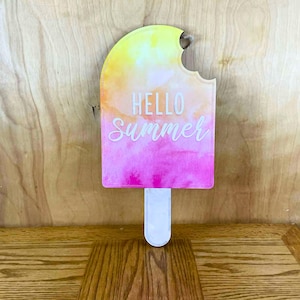 May include: A colorful popsicle-shaped sign with a bite taken out of the top. The sign features a watercolor design with yellow, orange, and pink hues. The words "HELLO Summer" are written in white script across the front. A white stick extends from the bottom.