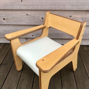 May include: A light wood chair with a white cushion. The chair has a simple, modern design with a high back and wide arms.