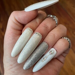 Puede incluir: Uñas acrílicas largas, blancas, de forma ovalada con purpurina plateada y pedrería. Algunas uñas tienen un diseño de remolino blanco.