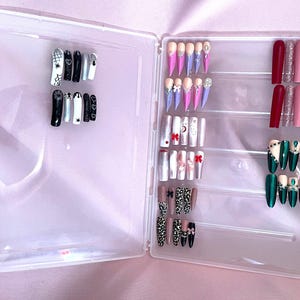 May include: A clear plastic case, open to display various sets of artificial nails. Designs include spiderwebs, moons, bows, leopard print, and glitter. Colors include black, white, pink, red, green, and nude. The nails are in various shapes, including coffin and stiletto.