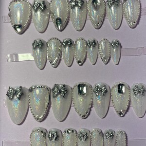 Peut inclure: Un ensemble de faux ongles blancs irisés, ornés de nœuds argentés et de strass. Les ongles de forme ovale sont disposés en rangées dans une boîte violette, présentant un design de nail art glamour.