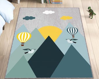 Puede incluir: Una alfombra gris y azul con una escena de montaña. La alfombra presenta un sol amarillo, dos globos aerostáticos y dos pájaros volando sobre las montañas. La alfombra tiene un fondo gris claro con nubes blancas.