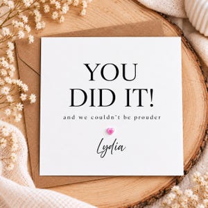 Peut inclure: Une carte de vœux carrée blanche avec le texte noir YOU DID IT! and we couldn't be prouder. Sous le texte se trouve une petite décoration en forme de cœur rose et le prénom Lydia écrit en police cursive. La carte est sur une enveloppe brune.