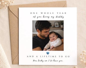 May include: A square greeting card with a white background and a central photograph of a man holding a baby. The text reads: ONE WHOLE YEAR of you being my daddy, AND A LIFETIME TO GO, How lucky am I to have you. A small blue heart is centred.