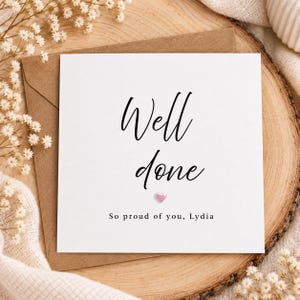 May include: A square white greeting card with black text that reads Well done in a script font, followed by a small pink heart icon and the text So proud of you, Lydia in a simple sans-serif font, placed on a brown paper envelope.
