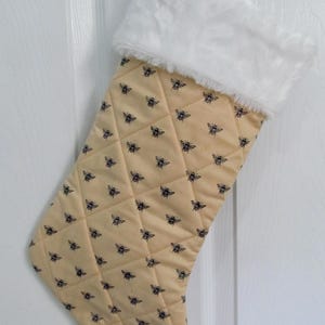May include: A quilted Christmas stocking with a cream-colored background and a pattern of black bees. The top of the stocking is trimmed with white faux fur. A white loop is attached for hanging.