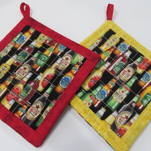 May include: Two quilted pot holders with a black background and a pattern of beer bottles. One pot holder has a red border and the other has a yellow border.