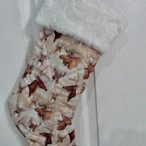 May include: A white faux fur-trimmed Christmas stocking with a quilted design featuring a pattern of brown deer heads.