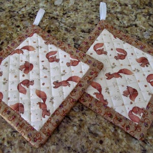 May include: Two square quilted pot holders with a white background featuring a pattern of brown squirrels and foliage. The edges are trimmed with a brown fabric with a floral design. Each pot holder has a white loop for hanging.