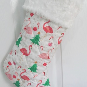 May include: A white Christmas stocking with a faux fur cuff. The stocking is decorated with a pink flamingo pattern, green Christmas trees, candy canes, and red gift boxes.