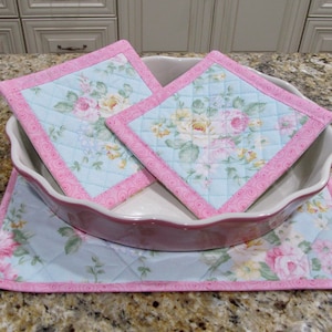 Puede incluir: Tres posavasos acolchados con un patrón floral en tonos de rosa, amarillo y azul. Los posavasos tienen un borde rosa con un diseño de espiral. Los posavasos están encima de una fuente para hornear de cerámica blanca.