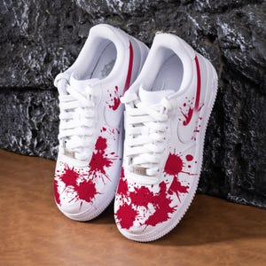 May include: White trainers with a red splatter paint design. The shoes feature white laces, a white sole, and a red stripe down the back. The Nike swoosh is visible on the side.