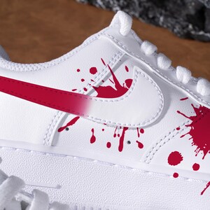 May include: Close-up of a white trainer with red detailing. The shoe has a red swoosh and red paint splatter designs. White laces and stitching are visible on the shoe.