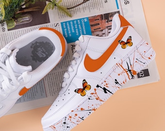 May include: White sneakers with orange accents and butterfly decals. The shoes feature an orange Nike swoosh and paint splatter details. The sneakers are on a newspaper background.