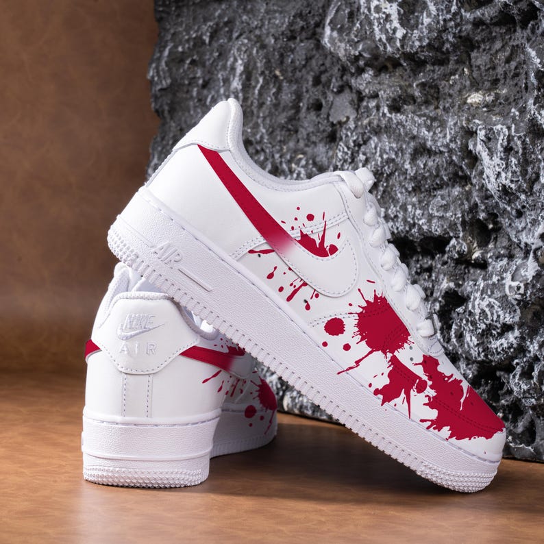 May include: White trainers with red detailing and a splatter design. The shoes feature the Nike logo and a red swoosh. The design includes red paint splatters across the white leather.