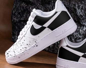 Può includere: Sneakers Nike Air Force 1 bianche e nere con un swoosh nero e un design a spruzzo di vernice nera. Le sneakers hanno un'intersuola bianca e una suola esterna nera.