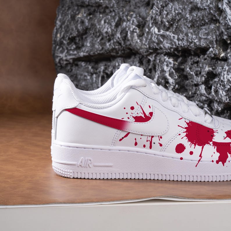 May include: White leather trainers with red detailing and a splattered paint design. The trainers have a red swoosh and red paint splatters on the side. The word "AIR" is visible on the midsole.