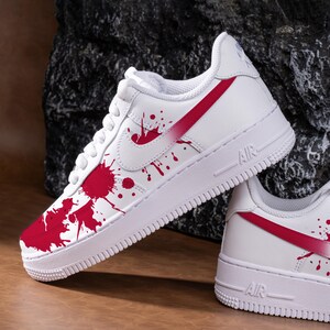May include: White trainers with a red splatter paint design. The shoes feature a red swoosh and a red stripe on the side. The soles are white with the word "AIR" printed on the side.