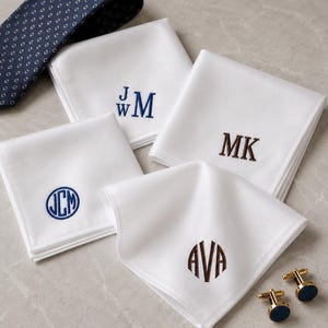 May include: Four white square pocket squares with embroidered monograms in blue and brown thread, including JWM, MK, JCM, and AVA. A dark blue patterned necktie and two gold-toned cufflinks with dark blue circular inlays are also visible.