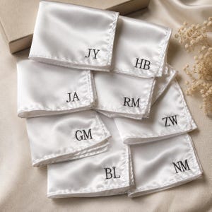 Może przedstawiać: Kolekcja białych satynowych poszetek, każda z wyhaftowanymi czarnymi inicjałami. Widoczne monogramy to JY, HB, JA, RM, ZW, GM, BL oraz NM. Te prostokątne kawałki materiału mają gładkie, odblaskowe wykończenie i są ułożone w nakładający się stos.
