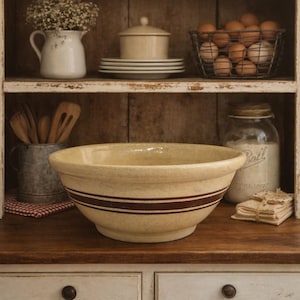 May include: A large, cream-colored stoneware mixing bowl with two dark brown stripes. The bowl sits on a wooden surface in a rustic kitchen setting, alongside other vintage kitchenware, including a wire basket of eggs and a jar of flour.