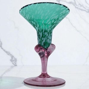 May include: A decorative glass vase with a flared top and a round base. The vase features a vibrant green speckled design on the top, transitioning to a pink stem and base. The vase is approximately 10 inches tall.