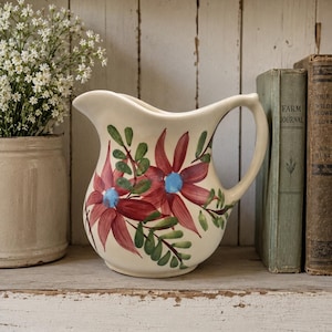 May include: A cream-colored ceramic pitcher featuring a hand-painted floral design with large red petals, bright blue centers, and green leaves. The pitcher has a curved handle and a wide pouring spout, showcasing a smooth, glossy finish throughout.